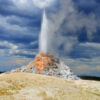 White Dome Geyser Erupts In Low Light Under A Dark Cloud Deck On A Rainy Day Over The Lower Geyser Basin Of Yellowstone National Park, Wyoming